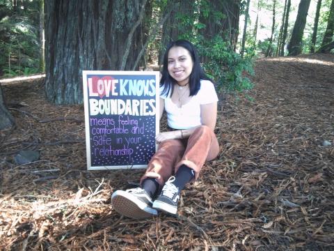 Feminine-presenting individual sitting on the forest floor and holding up chalkboard that says "Love knows boundaries means feeling comfortable and safe in your relationship." 